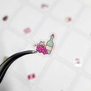 May include: A small sticker featuring a green wine bottle with a red label and a cluster of purple grapes. The sticker is held by a pair of tweezers against a white background, suggesting a crafting or scrapbooking theme.