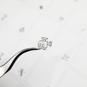 May include: A small, transparent sticker with a black outline of a meat grinder. The sticker is held by silver tweezers against a white background with faint, abstract shapes.