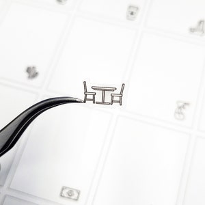 Könnte beinhalten: Ein kleiner, transparenter Aufkleber mit einem Tisch und zwei Stühlen, alle schwarz umrandet. Der Aufkleber wird von einer Pinzette vor einem weißen Hintergrund mit anderen kleinen, schwachen Aufkleberdesigns gehalten.