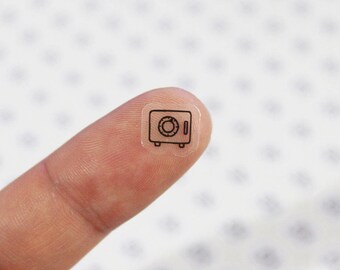 May include: A small, transparent sticker featuring a black outline of a safe. The safe has a circular dial and a rectangular door. The sticker is held on a finger, showcasing its small size and detail.