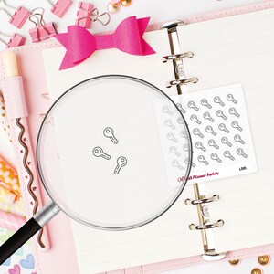 Può includere: Un'agenda rosa è aperta su una pagina a righe con tre piccoli adesivi neri a forma di chiave. Un foglio di adesivi a forma di chiave è visibile, insieme a un fiocco rosa, graffette e altri articoli di cancelleria. Gli adesivi sono di Olivia Planner Factory.