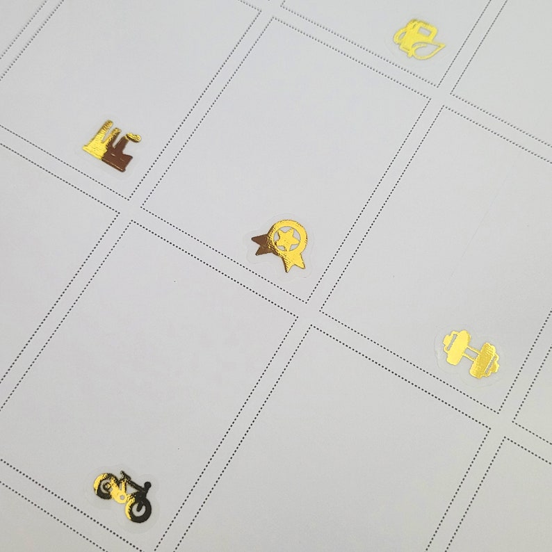 May include: A white grid with various gold foil stickers. The stickers include a factory, a star-shaped award ribbon, a motorcycle, a dumbbell, and a tea kettle. The grid is divided by dotted lines.