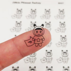 Peut inclure: Un autocollant transparent représentant un personnage de dessin animé avec une expression étourdie, portant une tenue à capuche avec une étoile et tenant une bannière avec la lettre "F". L'autocollant est tenu sur un doigt, avec d'autres autocollants similaires en arrière-plan.