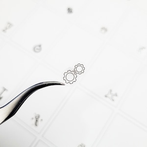May include: A pair of small, transparent stickers featuring a black outline of two interlocking gears. The stickers are held by a silver tweezer against a white background with faint, scattered illustrations.
