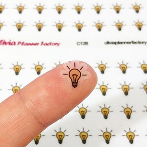 May include: A clear sticker featuring a brown light bulb illustration. The sticker is on a fingertip, with numerous other light bulb stickers visible in the background.