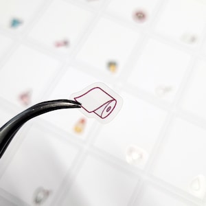 Può includere: Un adesivo trasparente con un contorno rosa di un rotolo di carta igienica. L'adesivo è tenuto da una pinzetta.