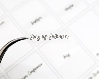 May include: A clear plastic label with the text "Song of Solomon" written in brown cursive script. The label is being held by tweezers.