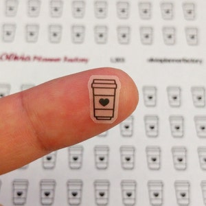 May include: A sheet of clear stickers with a black outline of a coffee cup with a heart inside. The stickers are small and are being held by a finger.