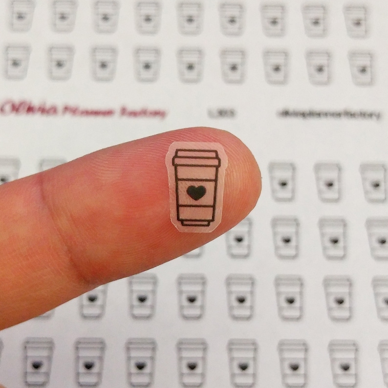 May include: A sheet of clear stickers with a black outline of a coffee cup with a heart inside. The stickers are small and are being held by a finger.