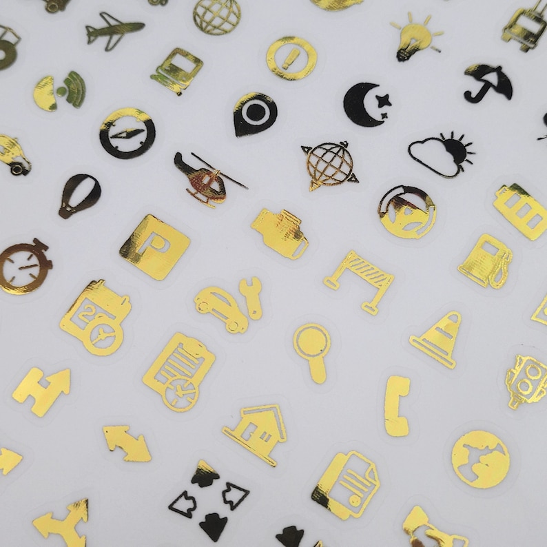 May include: A selection of gold foil stickers with travel and transport-themed icons. The stickers depict aeroplanes, cars, trains, compasses, and other related symbols. The stickers are presented on a white background.