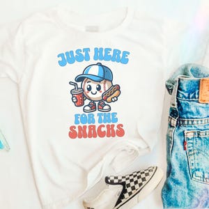 May include: White t-shirt with a cartoon baseball character holding a hot dog and a soda. The text on the shirt reads "Just Here For The Snacks".