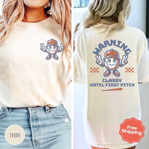 May include: A white t-shirt with a baseball character graphic on the front and the text "WARNING CLASSY UNTIL FIRST PITCH" on the back. The shirt is shown on a model wearing blue jeans.