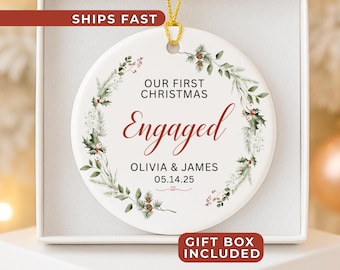 Peut inclure: Ornement de Noël en céramique blanche avec une ficelle dorée, portant l'inscription "OUR FIRST CHRISTMAS ENGAGED" en script rouge. Les noms Olivia & James et la date 05.14.25 sont en dessous. L'ornement est décoré d'une couronne florale verte et rouge.