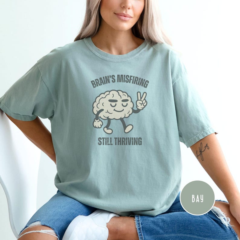 Pu&ograve; includere: T-shirt verde chiaro con la scritta "Brain's Misfiring Still Thriving" e un cervello dei cartoni animati che fa il segno della pace. La maglietta &egrave; di vestibilit&agrave; ampia e ha maniche corte. Il colore &egrave; bay.