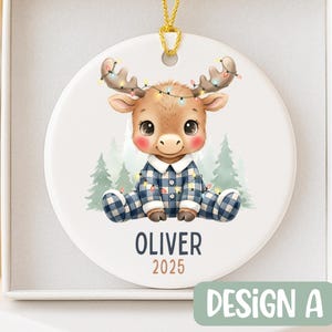 May include: A round Christmas ornament featuring a cartoon moose wearing blue plaid pajamas and a string of lights. The ornament has the name "OLIVER" and the year "2025" printed on it. The design is labeled "DESIGN A".