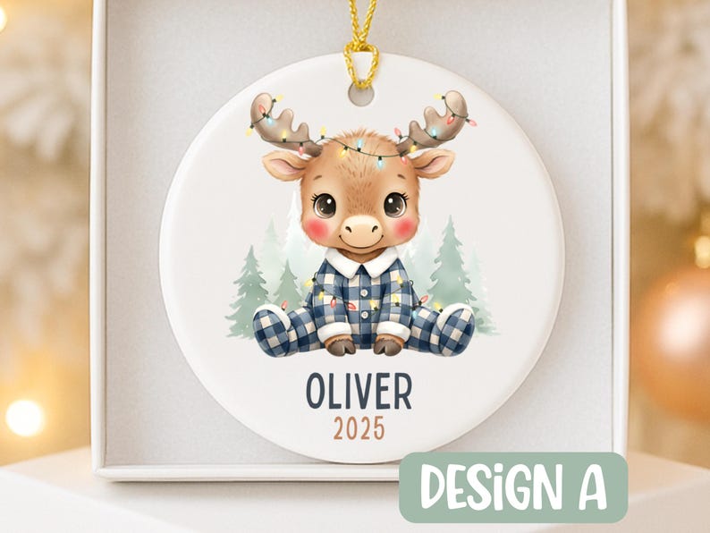 May include: A round Christmas ornament featuring a cartoon moose wearing blue plaid pajamas and a string of lights. The ornament has the name "OLIVER" and the year "2025" printed on it. The design is labeled "DESIGN A".