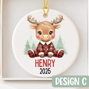May include: A round Christmas ornament featuring a cartoon moose wearing a red and black plaid onesie and a string of lights on its antlers. The name "HENRY" and the year "2025" are printed in red. The ornament is labeled "DESIGN C".