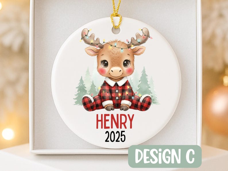 May include: A round Christmas ornament featuring a cartoon moose wearing a red and black plaid onesie and a string of lights on its antlers. The name "HENRY" and the year "2025" are printed in red. The ornament is labeled "DESIGN C".