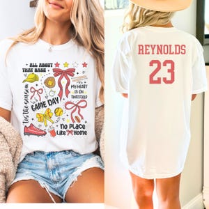 May include: White t-shirt with a red, white, and yellow baseball-themed design. The design includes a baseball, bats, a glove, a bow, and the text "Tis the season, Game Day, All About That Base, My Heart Is On That Field, No Place Like Home". The back of the shirt has the text "REYNOLDS 23" in red.