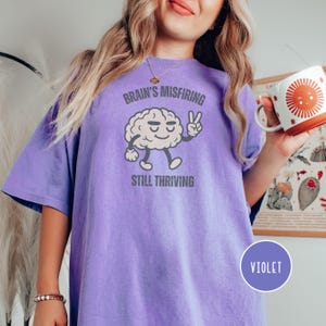 Pu&ograve; includere: T-shirt viola con la scritta "Brain's Misfiring Still Thriving" e un personaggio a forma di cervello dei cartoni animati che fa il segno della pace. La maglietta ha una vestibilit&agrave; ampia ed &egrave; realizzata in tessuto morbido.