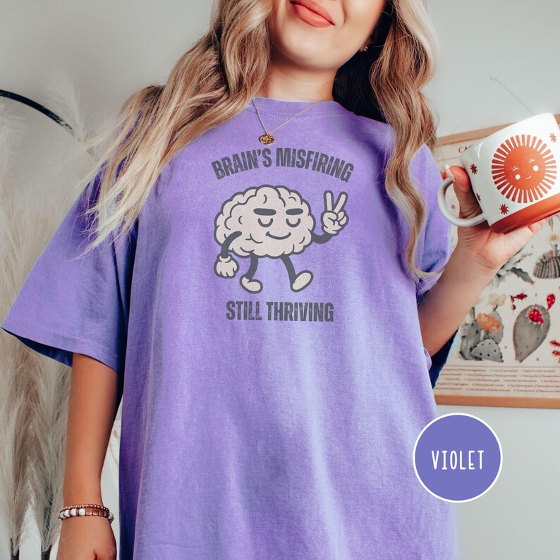 Pu&ograve; includere: T-shirt viola con la scritta "Brain's Misfiring Still Thriving" e un personaggio a forma di cervello dei cartoni animati che fa il segno della pace. La maglietta ha una vestibilit&agrave; ampia ed &egrave; realizzata in tessuto morbido.