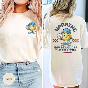 May include: A white t-shirt with a softball character graphic on the front and the text "WARNING MAY BE LOUDER THAN THE UMPIRE" on the back. The shirt is available in ivory.