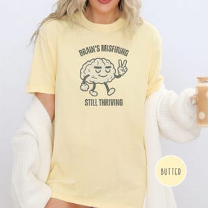 Pu&ograve; includere: T-shirt color burro con la scritta "Brain's Misfiring Still Thriving" e un personaggio a forma di cervello dei cartoni animati che fa il segno della pace. La maglietta &egrave; di colore giallo chiaro.