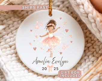May include: A white ceramic ornament featuring a ballerina illustration in a pink tutu, surrounded by stars and hearts. The ornament includes the name "Amelia Evelyn" and the numbers "20 25". A gold string is attached for hanging.