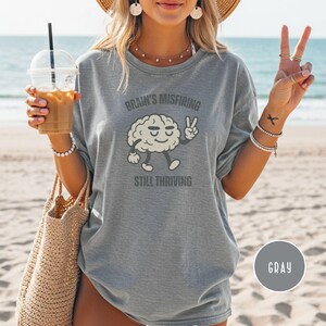 Pu&ograve; includere: T-shirt grigio con la scritta "Brain's Misfiring Still Thriving" e un personaggio a forma di cervello dei cartoni animati che fa il segno della pace. La maglietta &egrave; di vestibilit&agrave; ampia e ha maniche corte.