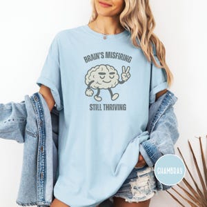 Pu&ograve; includere: T-shirt azzurra con la scritta "Brain's Misfiring Still Thriving" e un personaggio a forma di cervello dei cartoni animati che fa il segno della pace. La maglietta &egrave; realizzata in un materiale morbido e confortevole.
