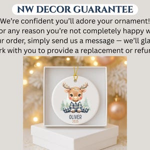 May include: A round ceramic ornament featuring a cartoon moose wearing a blue plaid outfit. The moose is sitting in front of two small green trees. The name "OLIVER" and the year "2025" are printed below the moose. The ornament is in a white box.