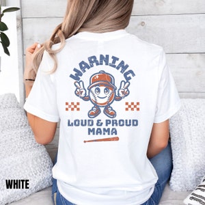 May include: White t-shirt with a distressed blue and orange baseball graphic. The graphic says "Warning" in a circle around a baseball with a face and a baseball cap. Below the graphic it says "Loud & Proud Mama" with a baseball bat underneath.