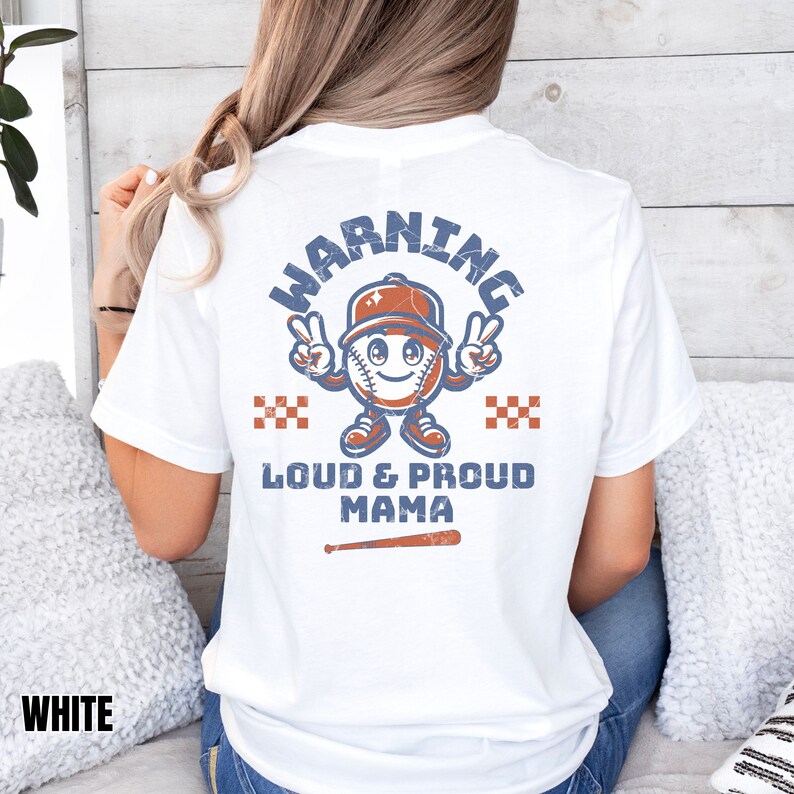 May include: White t-shirt with a distressed blue and orange baseball graphic. The graphic says "Warning" in a circle around a baseball with a face and a baseball cap. Below the graphic it says "Loud & Proud Mama" with a baseball bat underneath.