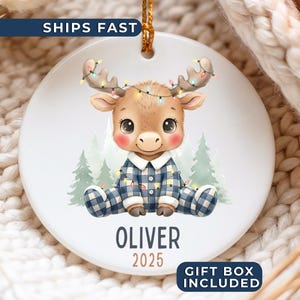 May include: A round ceramic Christmas ornament featuring a cartoon moose wearing blue plaid pajamas and a string of lights. The ornament has the name "Oliver" and the year "2025" printed on it. The ornament is white with a gold string for hanging.