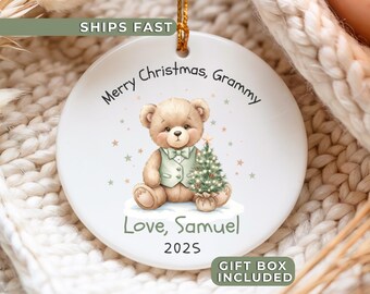 Peut inclure: Ornement de Noël rond et blanc avec une illustration d'ours en peluche. L'ours porte un gilet et tient un sapin de Noël. Le texte comprend "Merry Christmas, Grammy", "Love, Samuel" et "2025". Une ficelle dorée est attachée pour l'accrocher.