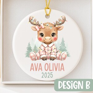 May include: A round Christmas ornament featuring a cartoon moose wearing pink pajamas and a bow. The moose is adorned with string lights and the ornament has the text "AVA OLIVIA 2025". The ornament is in a white box.