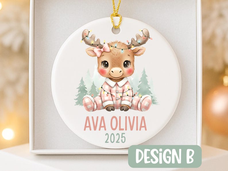 May include: A round Christmas ornament featuring a cartoon moose wearing pink pajamas and a bow. The moose is adorned with string lights and the ornament has the text "AVA OLIVIA 2025". The ornament is in a white box.