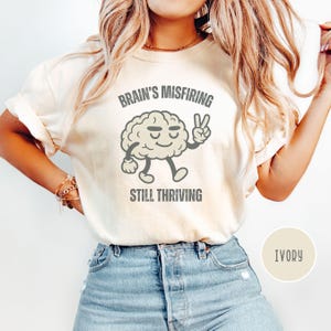 Pu&ograve; includere: T-shirt color avorio con la scritta "Brain's Misfiring Still Thriving" e un personaggio a forma di cervello dei cartoni animati che fa il segno della pace. La maglietta &egrave; indossata da una persona che indossa jeans blu.