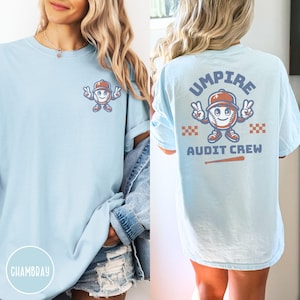 May include: Light blue long-sleeve t-shirt with a baseball-themed graphic on the front and back. The front features a baseball character giving peace signs. The back says "UMPIRE AUDIT CREW" with a baseball and bat graphic.