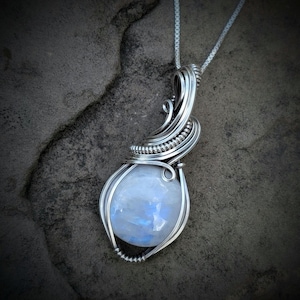 May include: A silver wire-wrapped pendant featuring a smooth, round, iridescent moonstone with blue flashes. The metal wire is intricately coiled around the stone and extends upward to form a bail, suspended from a thin silver box chain necklace.