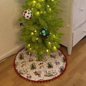 May include: A green artificial Christmas tree with white lights and red and green ornaments. The tree is decorated with candy canes and a red and white polka dot ornament. The tree is standing on a white and red tree skirt with a cartoon design.