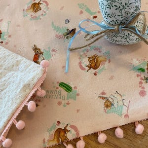 May include: A pink fabric with a repeating pattern of rabbits and squirrels, with a matching bunny-shaped decoration. The fabric has a border of pink pom-poms. A blue ribbon and twine are tied around the bunny decoration.
