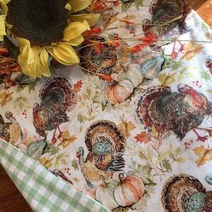 May include: A white table runner with a Thanksgiving theme featuring a repeating pattern of turkeys, pumpkins, gourds, and fall leaves. The runner has a green and white gingham border.