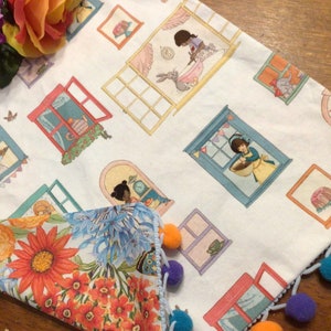 May include: White fabric with a colorful window pattern. Each window features a different scene with a person or animal. The fabric has a blue and white pom-pom trim. The fabric is folded over to reveal a floral pattern on the reverse side.