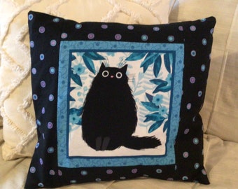 May include: Black and white pillow with a black cat sitting in a blue and white floral frame. The pillow has a black background with white polka dots and a blue border.