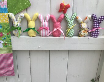 May include: A row of eight handmade Easter bunny decorations. The bunnies are made of fabric and are in various colors, including yellow, pink, green, and purple. They are all wearing a ribbon around their necks. The bunnies are sitting on a white wooden shelf.