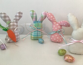 May include: Five handmade Easter bunny decorations. The bunnies are made of fabric and have different patterns, including plaid, gingham, and polka dots. Each bunny has a ribbon tied around its neck. There are also several Easter eggs scattered around the bunnies.