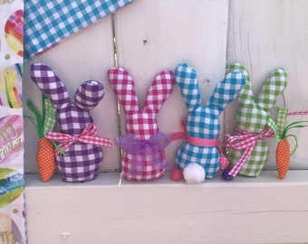 May include: Four gingham fabric Easter bunnies in purple, pink, blue, and green, each with a carrot and ribbon. The bunnies have white pom-pom tails and are arranged on a white surface. A fabric with Easter eggs and the words "Happy Easter" is on the left.