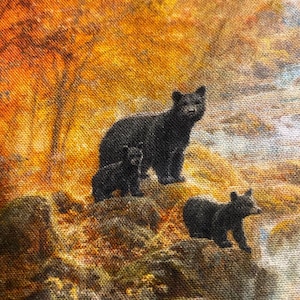May include: A fabric print featuring a black bear with two cubs in a natural setting. The bears are positioned on a rocky surface, with a backdrop of autumn foliage in shades of orange and yellow. The overall scene evokes a sense of wildlife and nature.