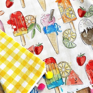 May include: A white fabric with a yellow and white gingham pattern on the left side. The fabric has a white trim with colourful pom-poms. The fabric is printed with a watercolour design of ice cream cones, popsicles, watermelon, strawberries, and lemons.
